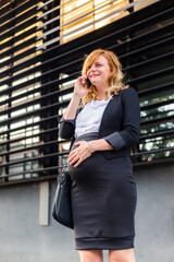 Fototapeta premium A smiling pregnant businesswoman standing in front of a modern building having a phone call.