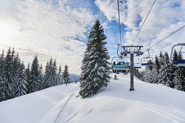 Panorama of snowy hills with coniferous trees, ski lifts and people under blue cloudy sky. Scenery of snow-covered slopes with chairlift mechanism for transporting skiers. Concept of ski resort.