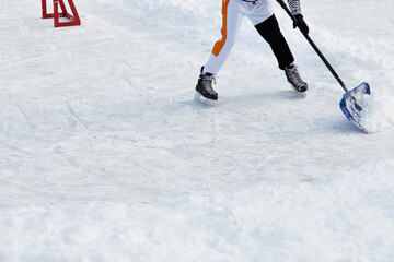 Hockey player on ice-skating removing snow with a blue shovel in the winter in a park. Wearing white clothes. Moving from goal to corner. Ice rink cleaning. Active sport outdoor.