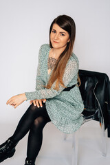 Frau sitz an Stuhl, grünes Blumenkleid, schwarze Lederjacke, braune lange Haare, feundlich, modern