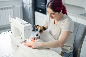 Caucasian woman sews while sitting in the kitchen. Dog Jack Russell Terrier sits on the lap of the owner. Home hobby.