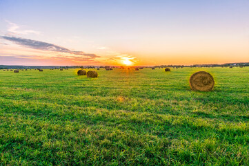 Scenic view at beautiful sunset in a green shiny field with hay stacks, cloudy sky, golden sun rays, anazing summer valley evening landscape