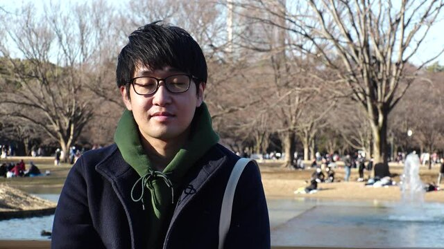 Asian (Japanese) young man sitting on the bench at the park in winter daytime. Smiling (shy smile), relaxing and looking at the camera. One person, late twenties, black hair with glasses.