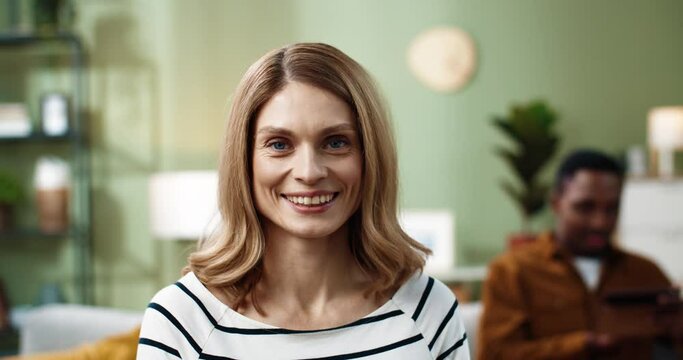 Close up portrait of happy caucasian woman sitting at home on background of busy man looking at camera smiling.
