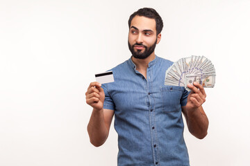 Satisfied positive man with beard looking away with cunning look holding money and credit card, choosing between cash and contactless payments. Indoor studio shot isolated on white background