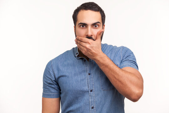 I will not say anything. Surprised embarrassed man in blue shirt closing mouth with hand looking at camera with confused expression, keeping silence. Indoor studio shot isolated on white background