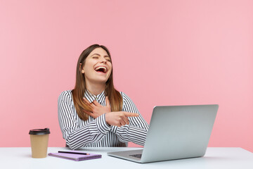 Funny positive brunette woman in striped shirt pointing to laptop screen and sincerely laughing talking on video call, sitting at workplace home office. Indoor studio shot isolated on pink background