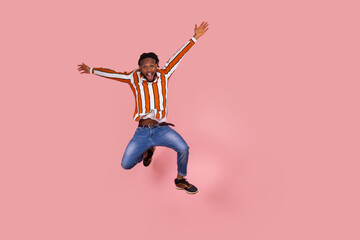 Fototapeta na wymiar Excited young man with dreadlocks highly jumping and rising hands, pretending he is flying, looking at camera with satisfied surprised expression. Indoor isolated on pink background