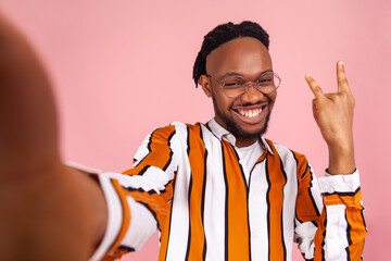 Happy smiling african man with dreadlocks in stylish striped shirt showing rock and roll gesture with fingers, posing at selfie camera, live stream. Indoor studio shot isolated on pink background