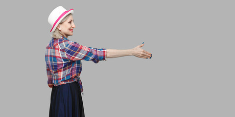 Profile side view portrait of happy modern stylish mature woman in casual style with hat standing, looking with toothy smile, giving hand to greeting. indoor studio shot isolated on gray background.