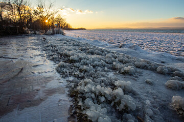 Frozen promenade on  a lake shore with sunset. Grass on the coast in a water frost. Ice covered plants, small halm and a freezing lake.