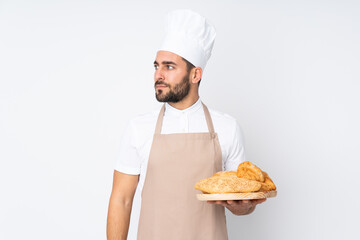 Male baker holding a table with several breads isolated on white background looking side
