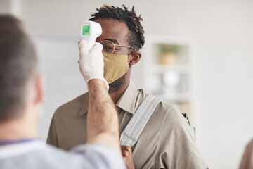 Shot of unrecognizable doctor checking temperature of young African American man wearing mask in...