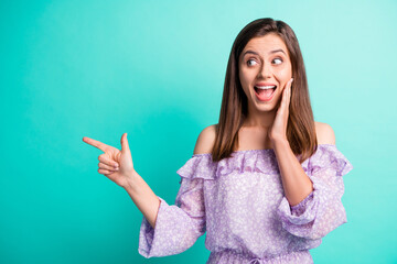 Fototapeta premium Portrait of impressed funky brunette girl pointing empty space hand face wear purple shirt isolated on blue color background