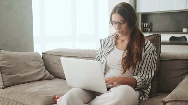 A focused concentrated young woman is sitting on the sofa and using her laptop at home