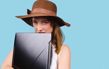 Portrait of a young blonde girl in a summer hat hugging a laptop on a blue background with empty side space for advertising.