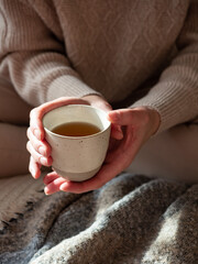 Light ceramic cup with black tea in the hands of a woman wearing a knitted beige woolen sweater in the morning sun