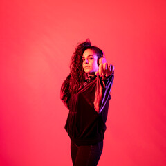 Vertical shot of a young girl with her brown curly hair in a ponytail wearing headphones pointing her hand to the camera, against red background