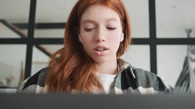 A focused redhead woman with earbuds is using her laptop computer sitting in the apartments