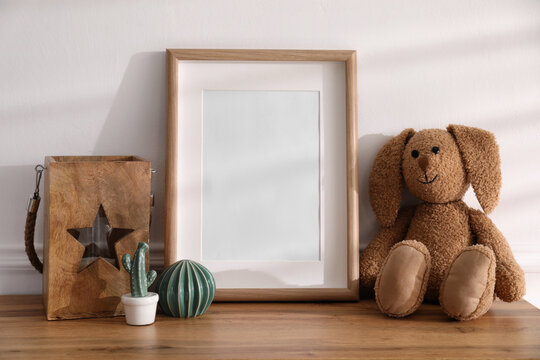 Empty photo frame near cute toy bunny and decor on wooden table, space for text. Baby room interior element