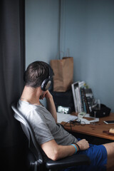 Lifestyle photo of adult young man in headphones sitting at the table with mobile home in front of him. Home office during pandemic coronavirus covid-19 concept. Internet and loneliness idea. 