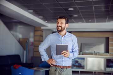 Smiling successful bearded chief standing in lobby of shipping firm, holding tablet and holding...
