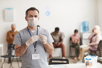 Waist up portrait of mature male doctor holding syringe and wearing mask while vaccinating patients in medical clinic, copy space