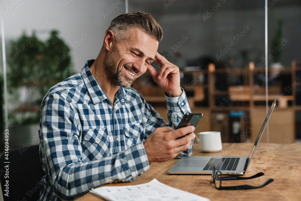Wall mural smiling grey man using mobile phone while working with laptop