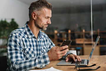 Focused grey man using mobile phone while working with laptop