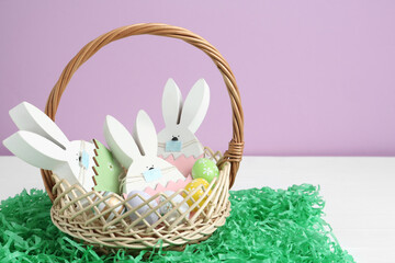 Wooden bunnies with protective masks, painted eggs in basket and paper grass on white wooden table. Easter holiday during COVID-19 quarantine