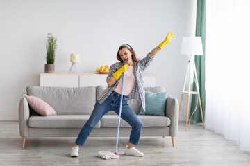 Happy young woman cleaning her home, singing at mop like at microphone and having fun, free space