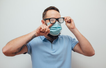 Handsome man with beard wiping blurred foggy misted glasses caused by wearing medical mask on light...