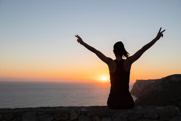 Backlit silhouette of a woman watching the sunrise over the sea making victory sign