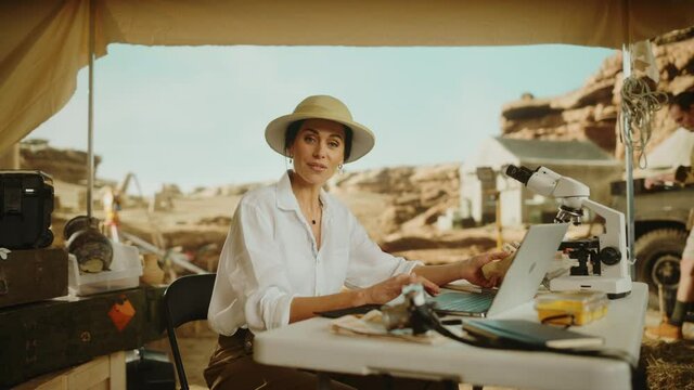 Archeological Digging Site: Portrait of Great Female Archaeologist Doing Research, Using Laptop, Looks at the Camera Smilingly while Holding Fossil Remains or Ancient Civilization Culture Artifacts