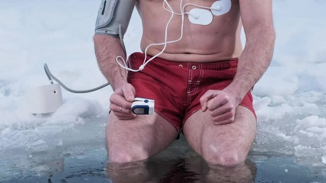 Medical test in the cold. Man shivers with cold while sitting on the ice with legs immersed into icy water. Oximeter, blood pressure monitor and cardio electrodes are connected to his body