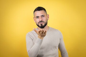 Handsome man with beard wearing sweater over yellow background looking at the camera blowing a kiss with hand on air being lovely and sexy. Love expression.
