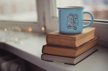 Closeup blue cup of coffee with vintage books on windowsill. Vintage light, blurred and bokeh background. Cosiness, warm and reading concept