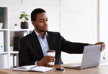 Fototapeta premium Young black businessman opening his notebook, drinking coffee, office interior