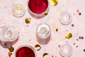 champagne flutes, wine glasses and vodka shots on a pink and gold paper confetti background
