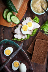 preparing sandwich with meat, dark bread, fresh green salad, eggs and cucumbers on rustic wooden table surface