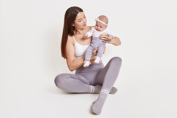 Attractive young woman wearing grey leggins and sleeveless t shirt sitting on floor with newborn child, dark haired female carries her daughter, kid with hairband, isolated on white wall.