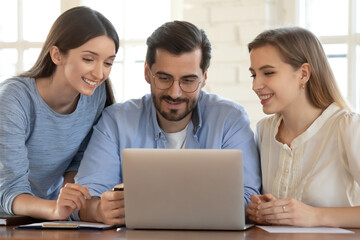 Smiling young male manager looking at computer screen with happy two female colleagues, discussing online project development, working brainstorming in office, using software or enjoying break time.