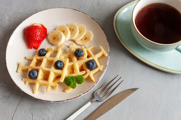 Close-up of soft Viennese waffles on the plate with blueberries, strawberry, chocolate sauce and cup of tea.