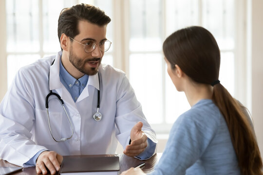 Pleasant young handsome male doctor in white uniform giving professional surgeon consultation to woman patient, explaining disease treatment protocol or discussing health condition at meeting.