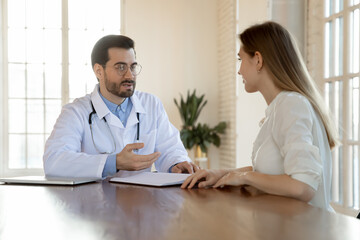 Concentrated young male general practitioner in eyewear and white coat giving professional medical consultation to female patient, discussing healthcare treatment or health test results at meeting.