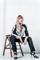 Fototapeta premium Blonde young woman with defiant eyes and loose hair on a low wooden stool. Model in studio on background of white brick wall in relaxed pose. Vertical image. Concept style, fashion, modeling.