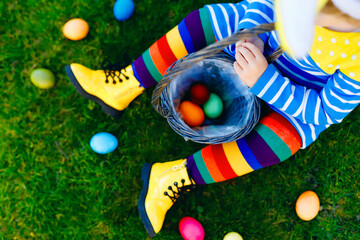 Close-up of legs of toddler girl with colorful stockings and shoes and basket with colored eggs. Child having fun with traditional Easter eggs hunt, outdoors. Unrecognizable face, no face.