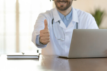 Close up smiling young male doctor showing thumbs up gesture, working on computer in clinic office, recommending distant online medical consultation to patient, medical insurance good service.