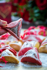 Hand showing pink colorful hamantash cookie with flowers and hamantaschen in background. Jewish traditional pastry for Purim (Jewish carnival holiday). Homemade gluten free cookie, chocolate filled.