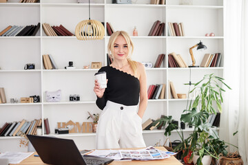 Young blond woman, wearing black top and white pants, standing by table with laptop, holding paper coffee cup. Office manager at her workplace. Female working in light office in company.